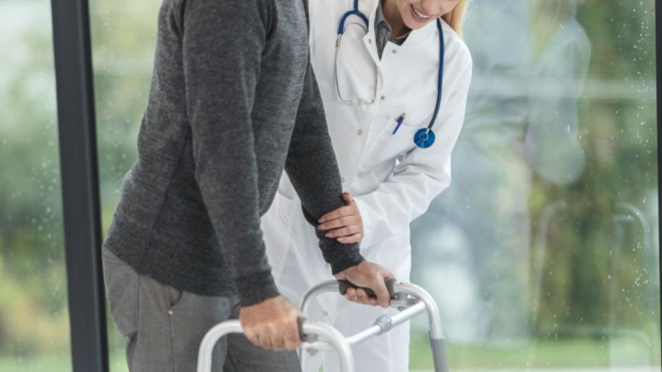 A care worker supporting an older man using a walking frame beside a window