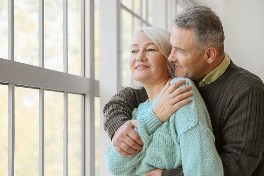 An older couple standing together by a bright window