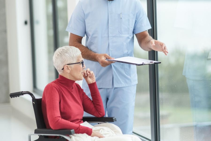 A care worker with a clipboard beside a seated resident in a bright room