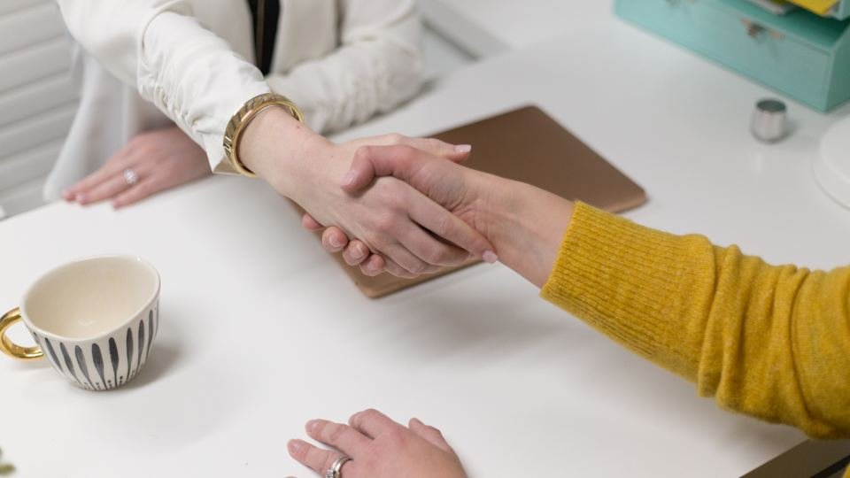 Two people shaking hands across a desk with paperwork between them