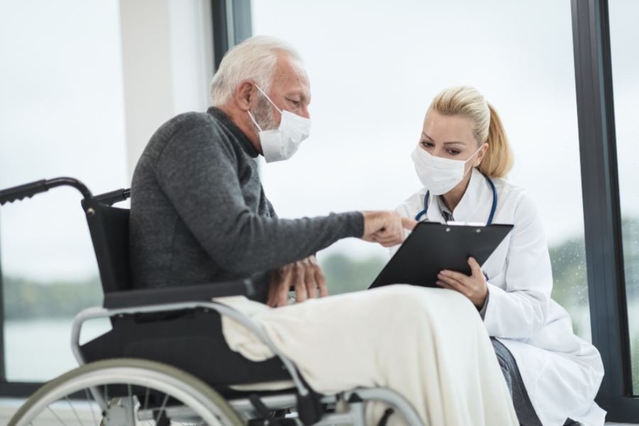 A nurse with a clipboard beside a seated older man in a care setting